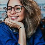 A young woman wearing glasses and denim, smiling in an indoor setting.